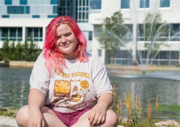 Rowan Armour, a person with pink hair and rainbow earrings, sits in front of Zumberge Pond. They are wearing a shirt that says &ldquo;Chosen Family.&rdquo; 