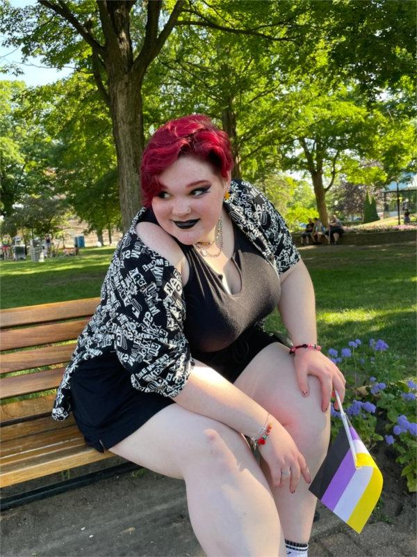 Zoey Belk, a person with short magenta hair, sits on a bench and holds a small non-binary flag with yellow, white, purple, and black stripes.