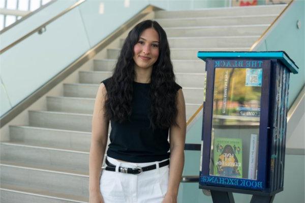 Jordyn Horton stands next to the Black Book Exchange Box in the Mary Idema Pew Library; staircase on right, box is painted deep blue